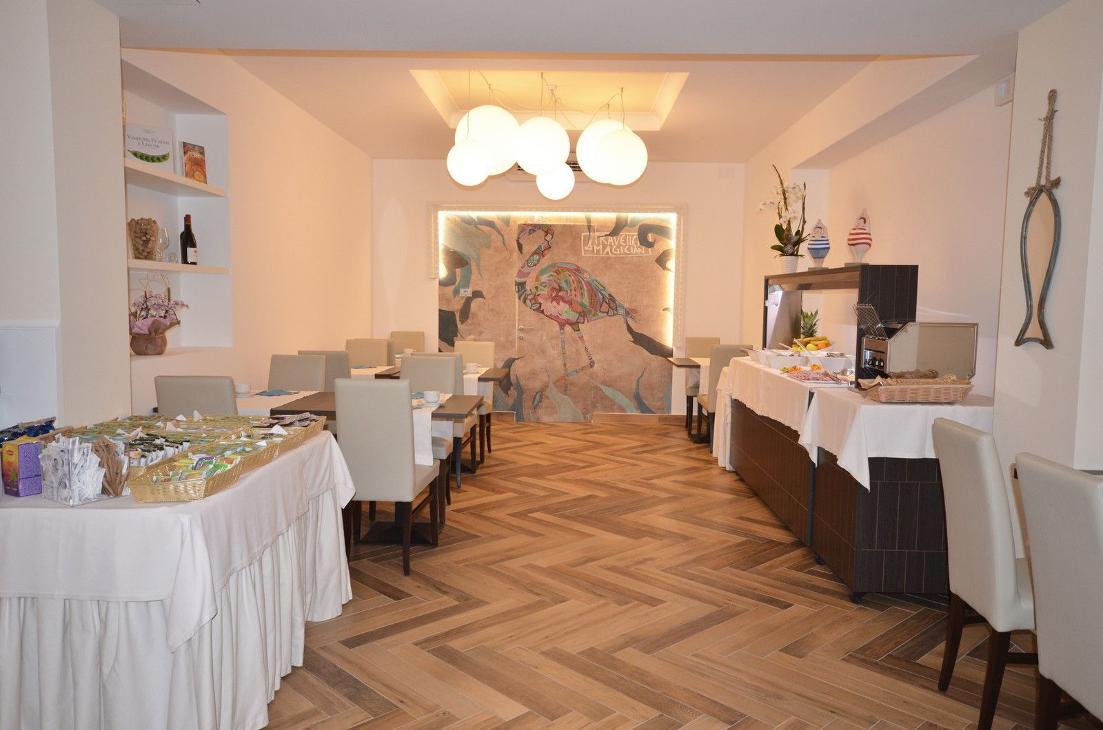 Ein Restaurant mit Holzdielenparkett, weißen Tischen und Stühlen. Es gibt eine Buffet-Ecke mit Speisen und Getränken, sowie eine Wand mit einem farbenfrohen Wandgemälde.