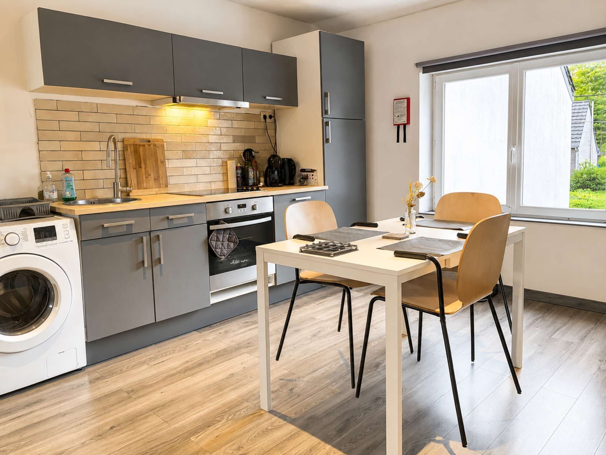 A modern kitchen with grey cabinets, a built-in oven, a washing machine, and a dining area with a table and chairs.