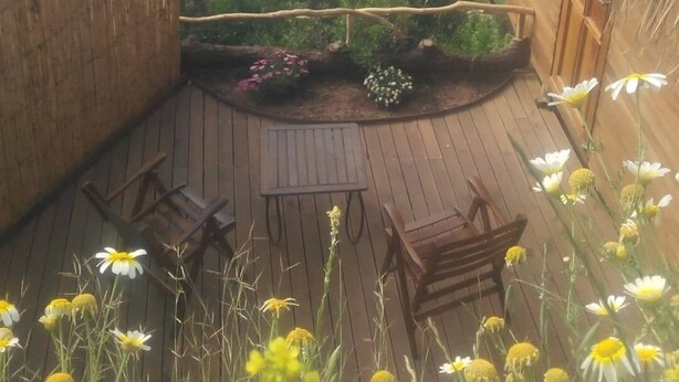 A wooden deck with a table and chairs, surrounded by a garden with flowers and a view of a turquoise sea. - Kök Kamp (Fethiye)