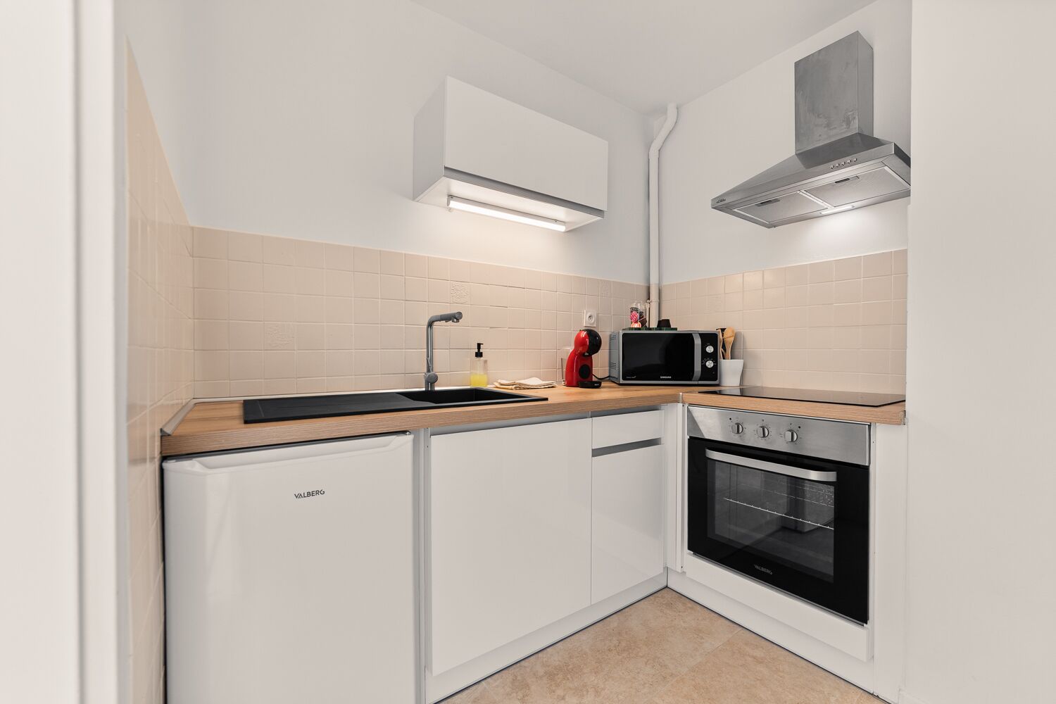 A compact kitchen with white cabinets, a black microwave, a built-in oven, and a stainless steel range hood.