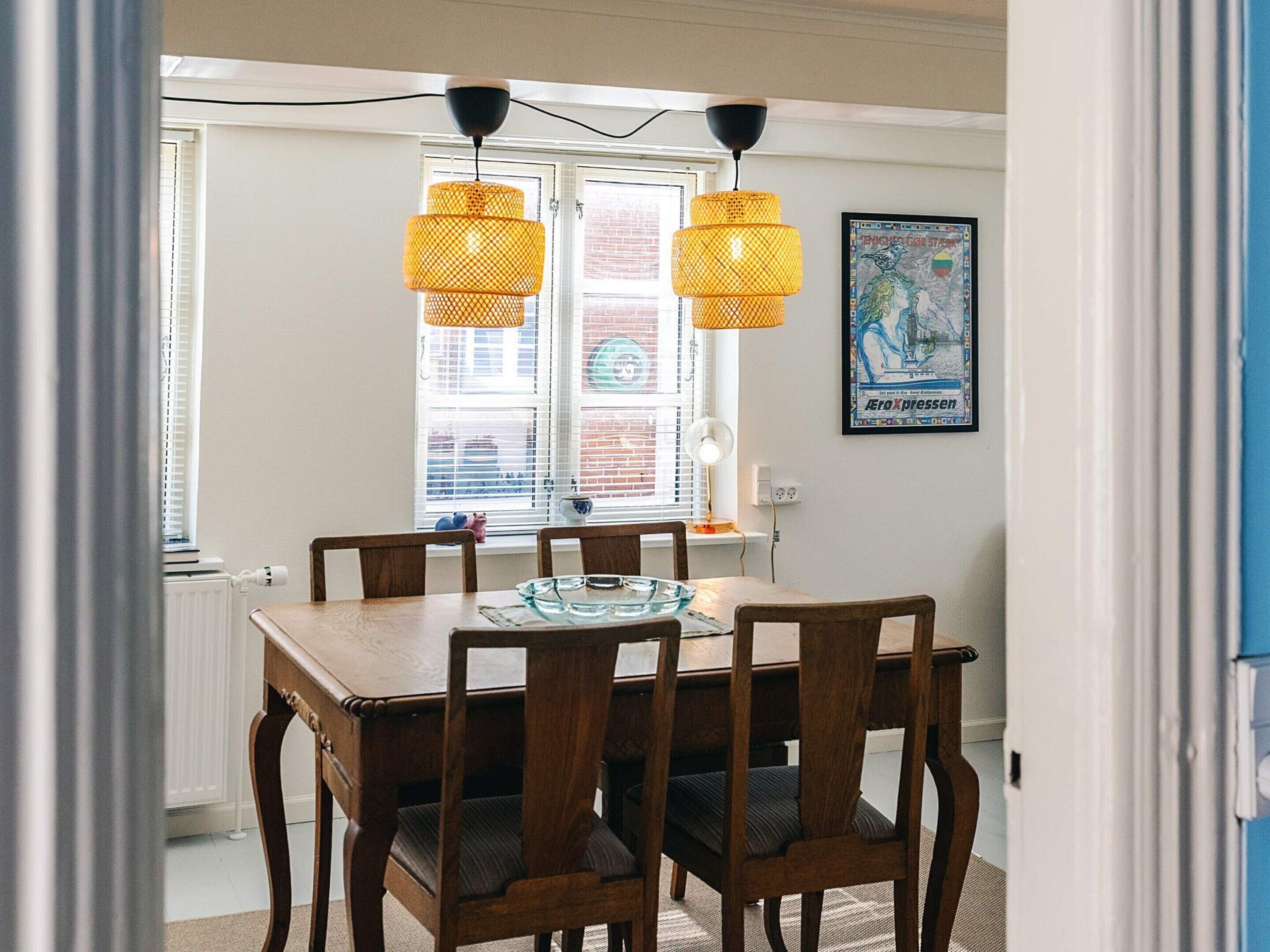 Un comedor con una mesa y sillas de madera, dos lámparas colgantes, una ventana con persianas y un cartel enmarcado en la pared.