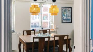 Un comedor con una mesa y sillas de madera, dos lámparas colgantes, una ventana con persianas y un cartel enmarcado en la pared.