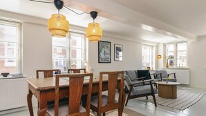 A spacious living room with a wooden dining table, grey upholstered chairs, and a grey sofa. There are two hanging lamps with a woven design, a framed map on the wall, and a radiator under the sofa. - 6 Person Holiday Home in Marstal (Marstal)