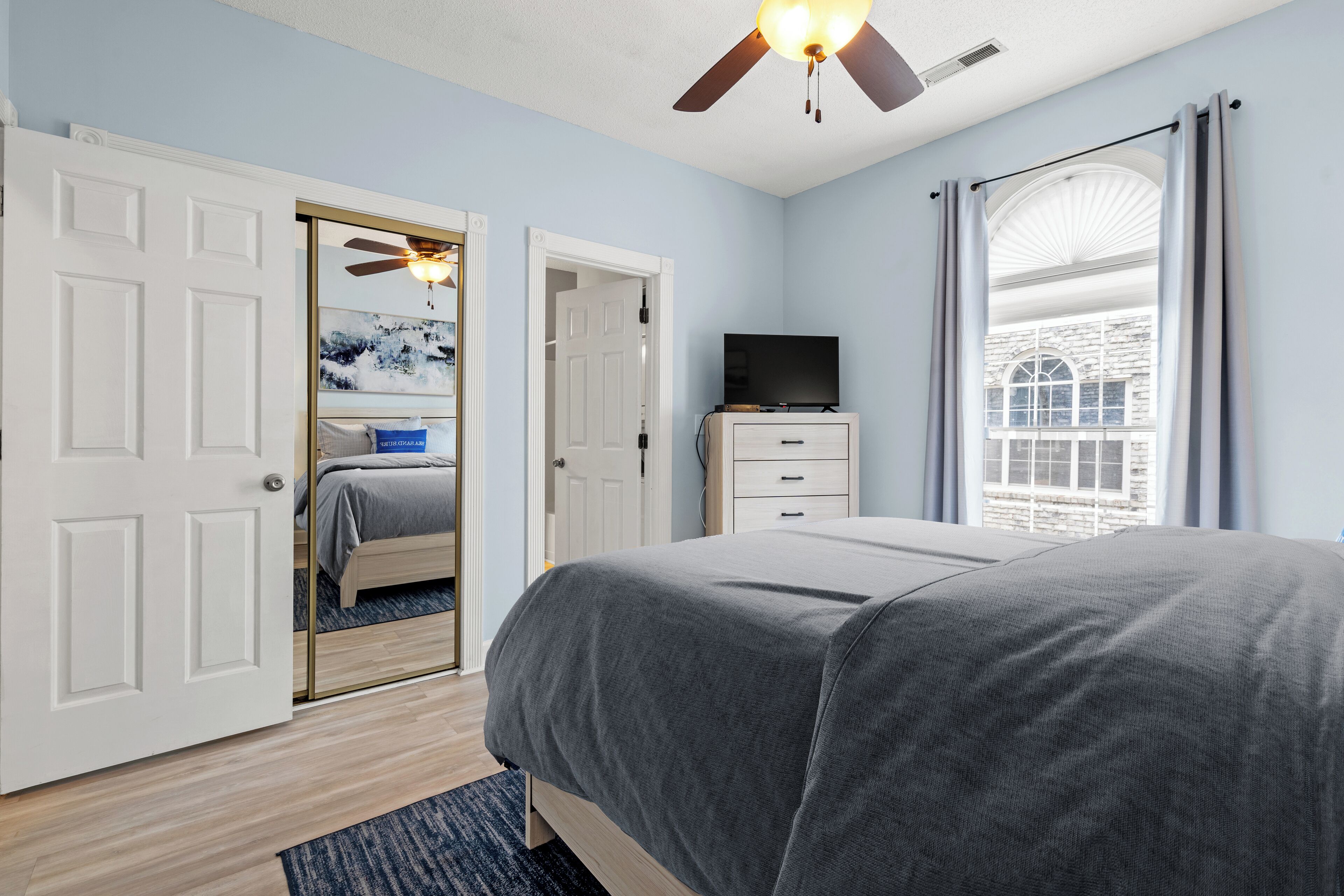 A bedroom with a bed, a television, a ceiling fan, and a window with curtains.