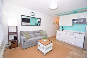 Een gezellig, ruimtelijk ingerichte leefruimte met een bank, een klein houten tafeltje, een kitchenette en een spiegel aan de muur.