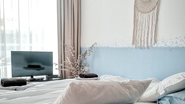 Un lit bien fait, avec une literie blanche et bleu clair, un téléviseur posé sur un meuble et une décoration murale en macramé.