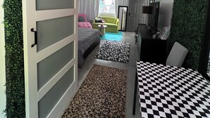 Un dormitorio con cama, escritorio y silla. Se ve una ventana con cortinas, una pared con una planta verde y un letrero de neón.
