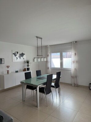 A modern dining area with a glass table, black chairs, a world map on the wall, and a window with curtains.