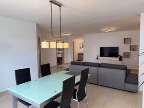 A modern living room with a glass-top dining table, grey sofa, and a flat-screen TV mounted on the wall.