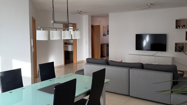 A modern living room with a glass dining table, black chairs, a grey sofa, and a flat-screen TV mounted on the wall. - Spacious apartment in the Palatinate Forest with terrace (Clausen)