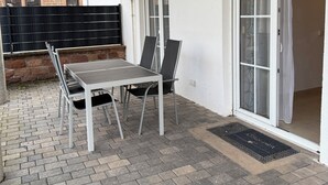 An outdoor patio with a table and chairs, a brick floor, and a white wall with a sliding glass door. - Spacious apartment in the Palatinate Forest with terrace (Clausen)