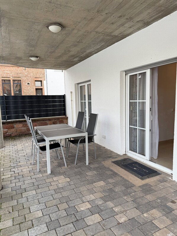 An outdoor patio with a table and chairs, a brick floor, and a white wall with a sliding glass door.