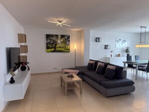 A modern living room with a grey sofa, a coffee table, and a flat-screen TV mounted on the wall. There is a dining area with a table and chairs, and a ceiling fan.