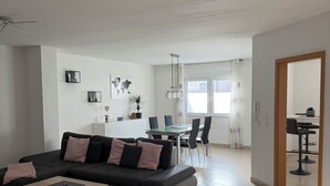 A modern living room with a dark grey sectional sofa, a wooden coffee table, and a dining area with a glass table and black chairs. - Spacious apartment in the Palatinate Forest with terrace (Clausen)