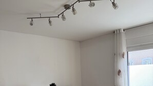 Une pièce au décor minimaliste, avec une table blanche et deux tabourets de bar noirs, un système d’éclairage sur rails au plafond et une fenêtre ornée d’un rideau blanc.