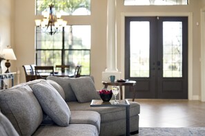 A living room with a grey sectional sofa, a small table with a bowl of fruit and a glass, and a chandelier.