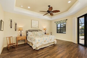 A bedroom with a large bed, two bedside tables with lamps, a ceiling fan, a window with a view, and a wooden chair. - Waterfront Haven | Pet Friendly | Heated Pool | Dock (Cape Coral)
