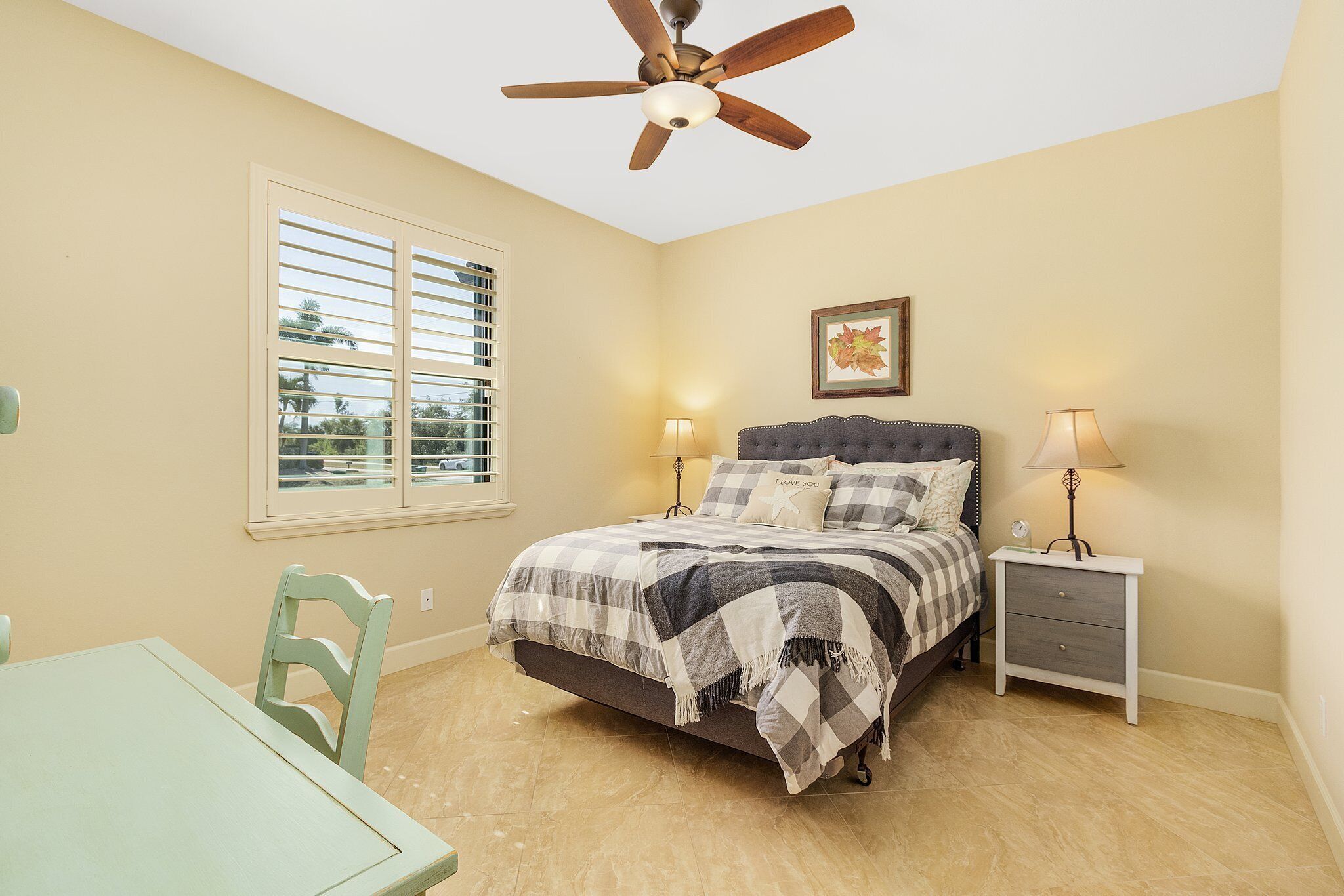 Quarto com cama, duas mesas de cabeceira com luminárias, um ventilador de teto, janela com persianas e uma cadeira.