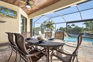 A covered outdoor dining area with wicker chairs and a table set for a meal, overlooking a pool and palm trees.