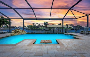 A swimming pool with a view of a sunset, surrounded by a covered area with chairs and a stone wall. - Waterfront Haven | Pet Friendly | Heated Pool | Dock (Cape Coral)