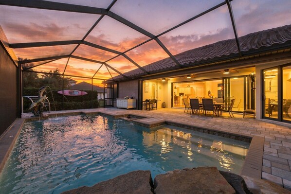 A swimming pool with a glass enclosure, a patio area with outdoor furniture, and a view of the sunset.