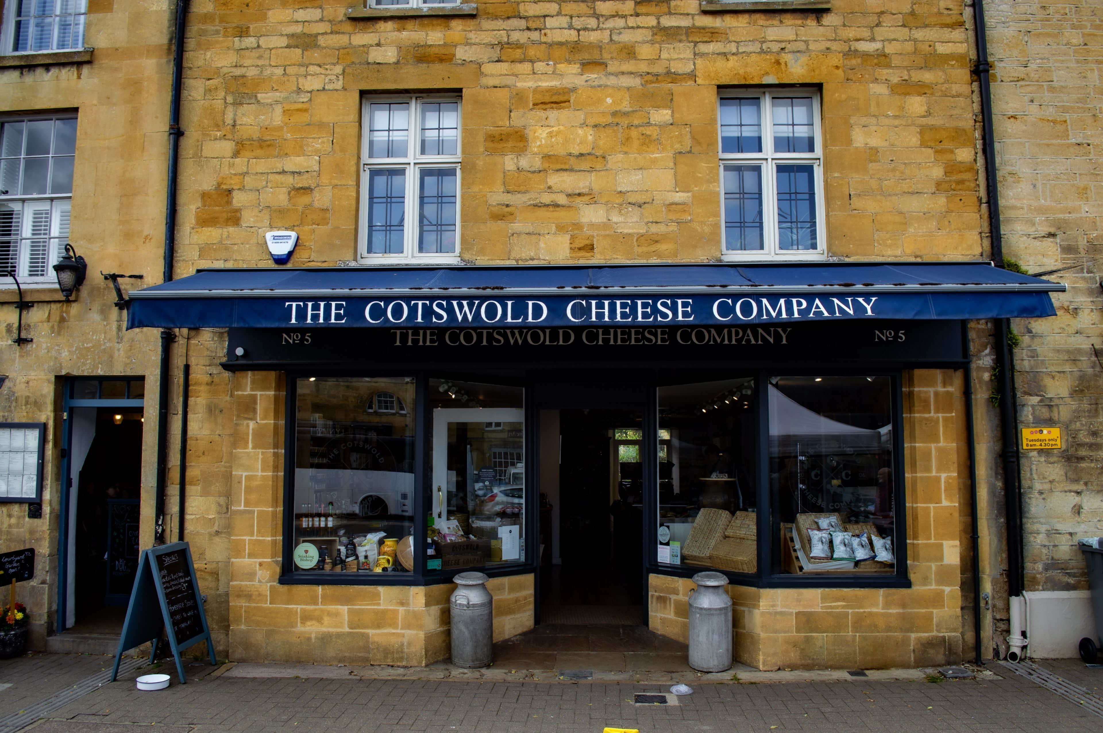 Un edificio de piedra con un toldo azul que dice 'The Cotswold Cheese Company'.