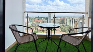 A balcony with two wicker chairs and a small round table, overlooking a cityscape with buildings and a clear sky. - One Bedroom Apartment in Kilimani, near Yaya Centre (Nairobi)