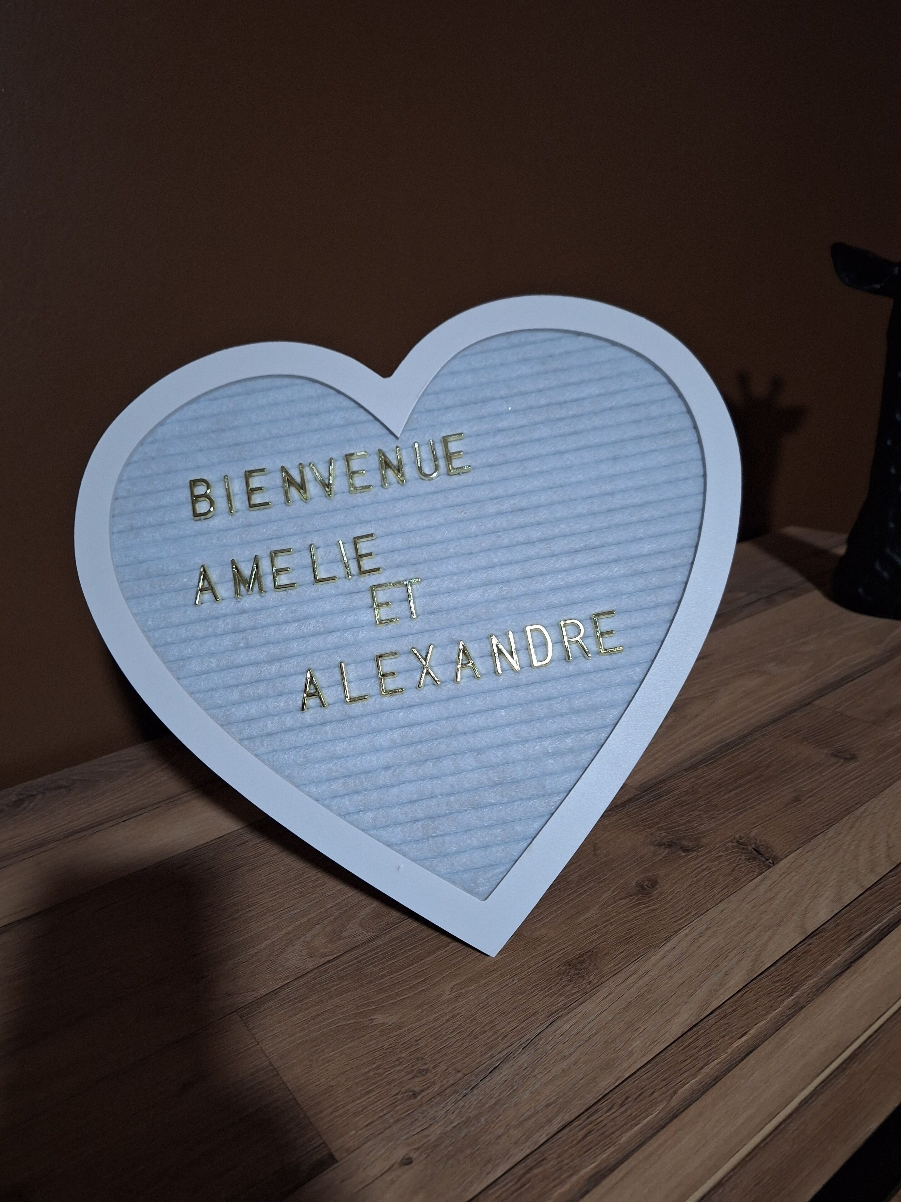 Ein herzförmiges Schild mit der Aufschrift "BIENVENUE AMELIE ET ALEXANDRE" auf einer Holzfläche.