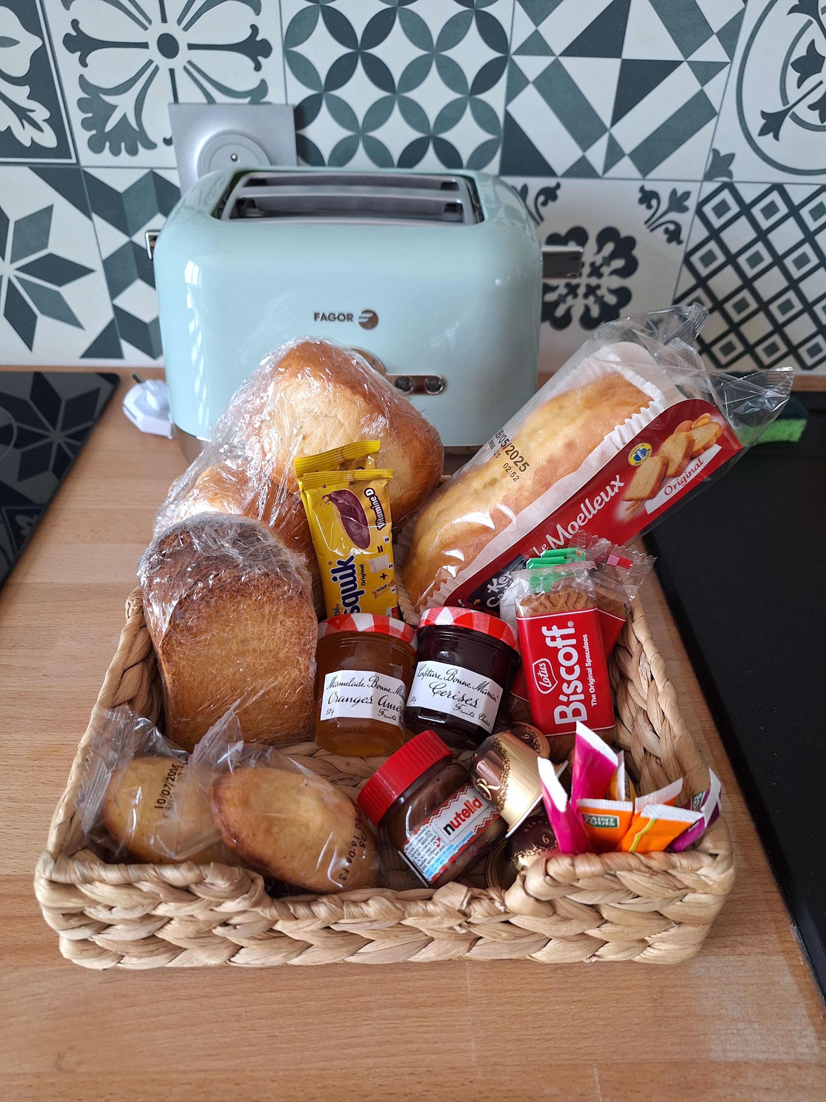 Rottingista punottu kori, joka on täynnä erilaisia ruoka-aineita, kuten leipää, hilloja ja keksejä. Kori on asetettu puupinnan päälle, ja taustalla näkyy leivänpaahdin.