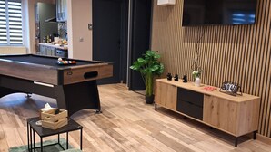 Een moderne woonkamer met een pooltafel, een groene bank, houten vloeren, een aan de muur bevestigde televisie en een houten kast met een klok.