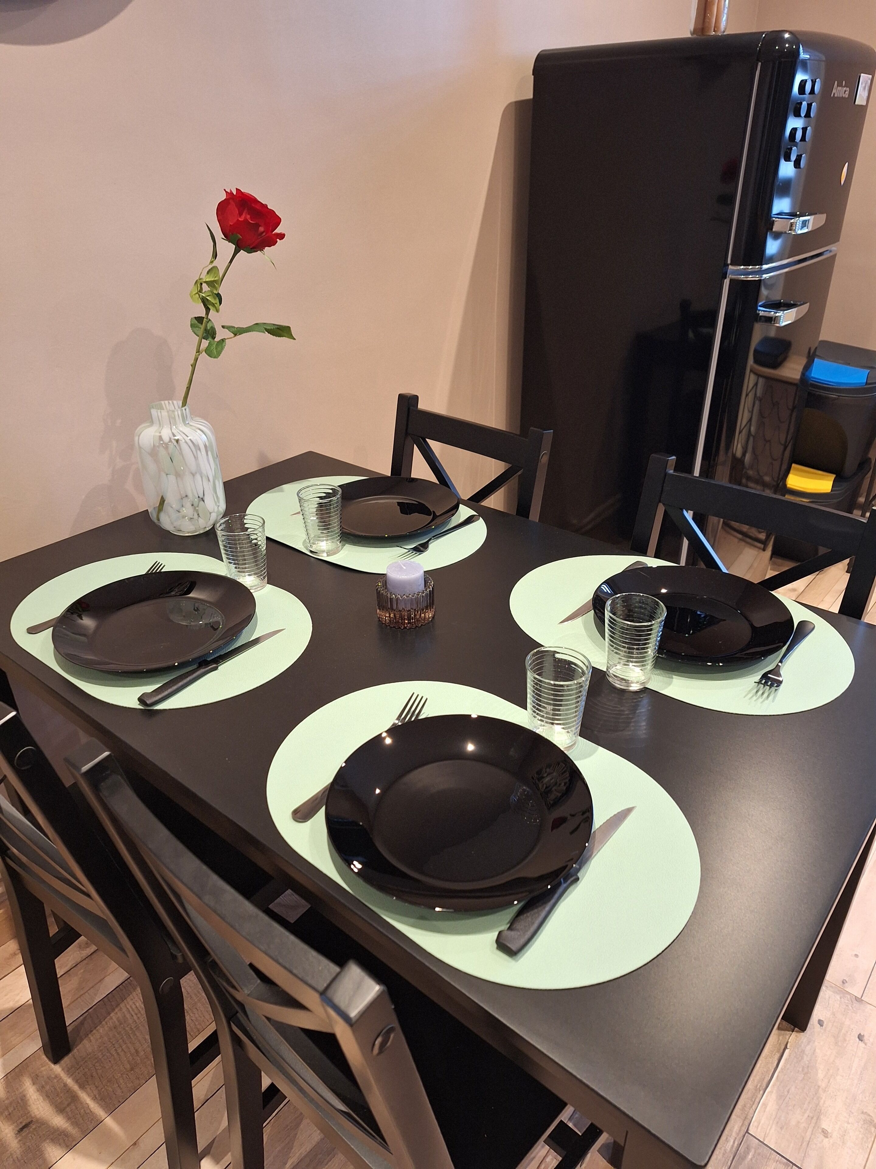 Tavola imbandita con piatti neri, posate e bicchieri, con un vaso di fiori bianchi e una rosa rossa a sinistra.