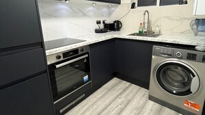 Cocina compacta con armarios oscuros, horno empotrado, lavadora y una ventana con persianas.