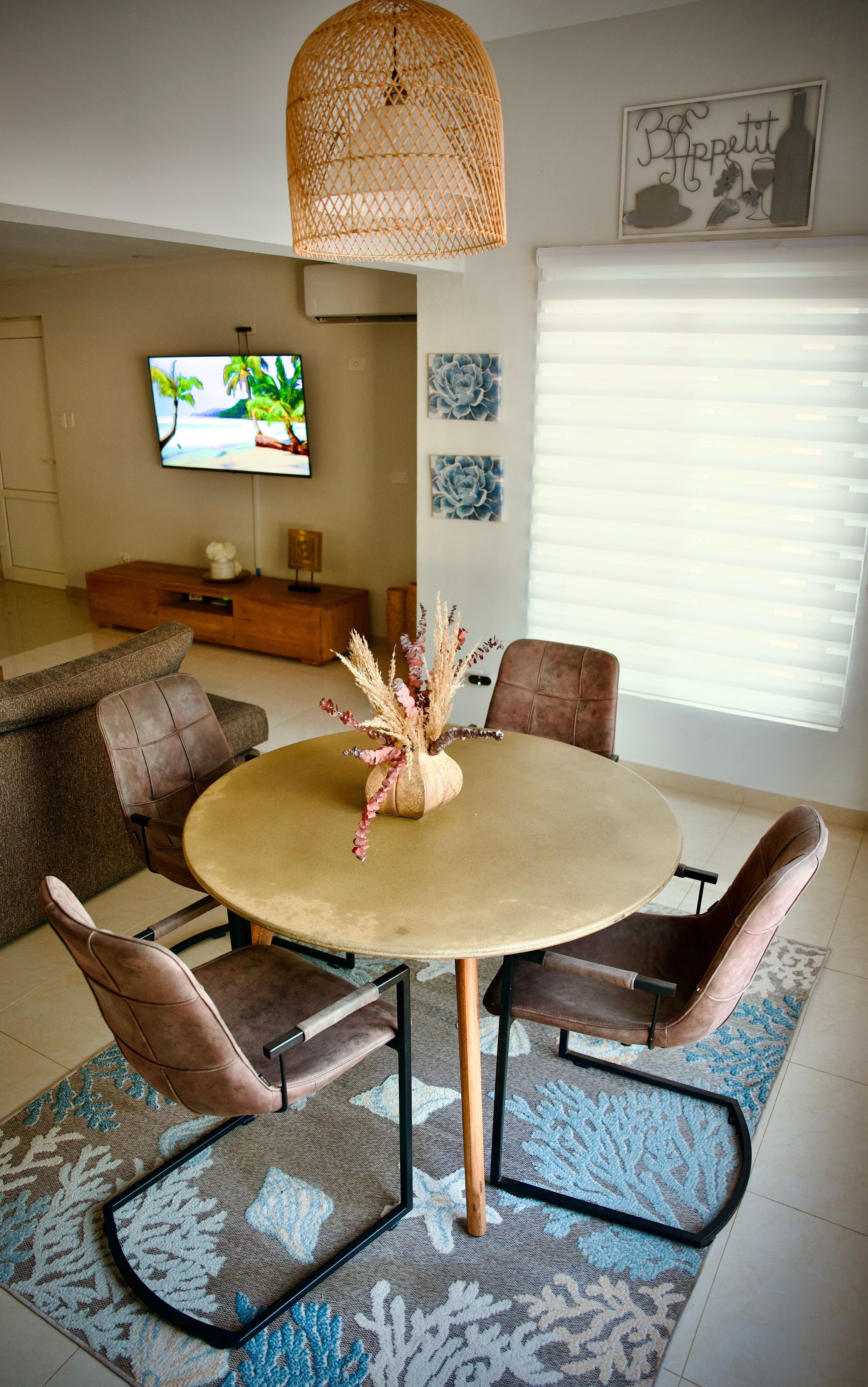 Uma sala de jantar com uma mesa redonda, quatro cadeiras, um vaso decorativo, uma televisão na parede e uma janela com persianas.