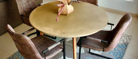 Uma sala de jantar com uma mesa redonda, quatro cadeiras, um vaso decorativo, uma televisão na parede e uma janela com persianas.