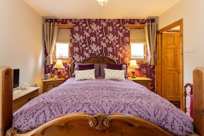 A bedroom with a large bed, two bedside lamps, a wooden headboard, and a floral-patterned wall. - Fishermans Cottage -  a cottage that sleeps 6 guests  in 3 bedrooms (Lossiemouth)