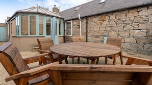Une table et des chaises en bois sont disposées à l’extérieur, avec un mur de pierre et une véranda en arrière-plan.