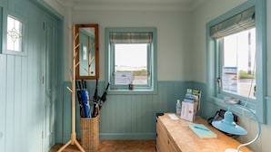 Une pièce aux murs de couleur turquoise, un bureau en bois avec une lampe, un porte-parapluie en osier et une fenêtre donnant sur la mer.