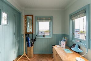A room with teal walls, a wooden desk with a lamp, a wicker umbrella stand, and a window with a view of the sea.