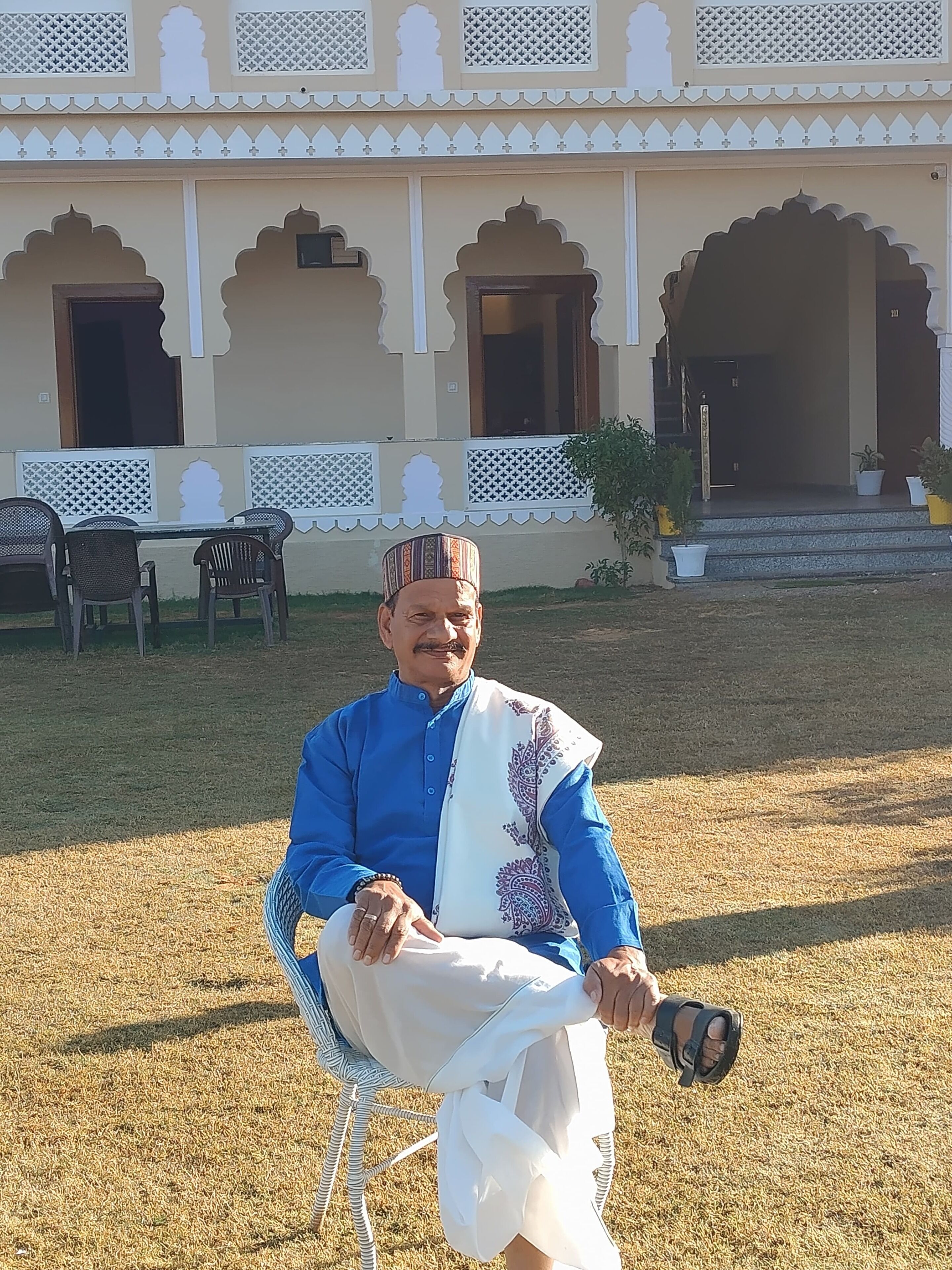 A man sitting on a chair in front of a building with traditional architecture, including arched doorways and decorative patterns.