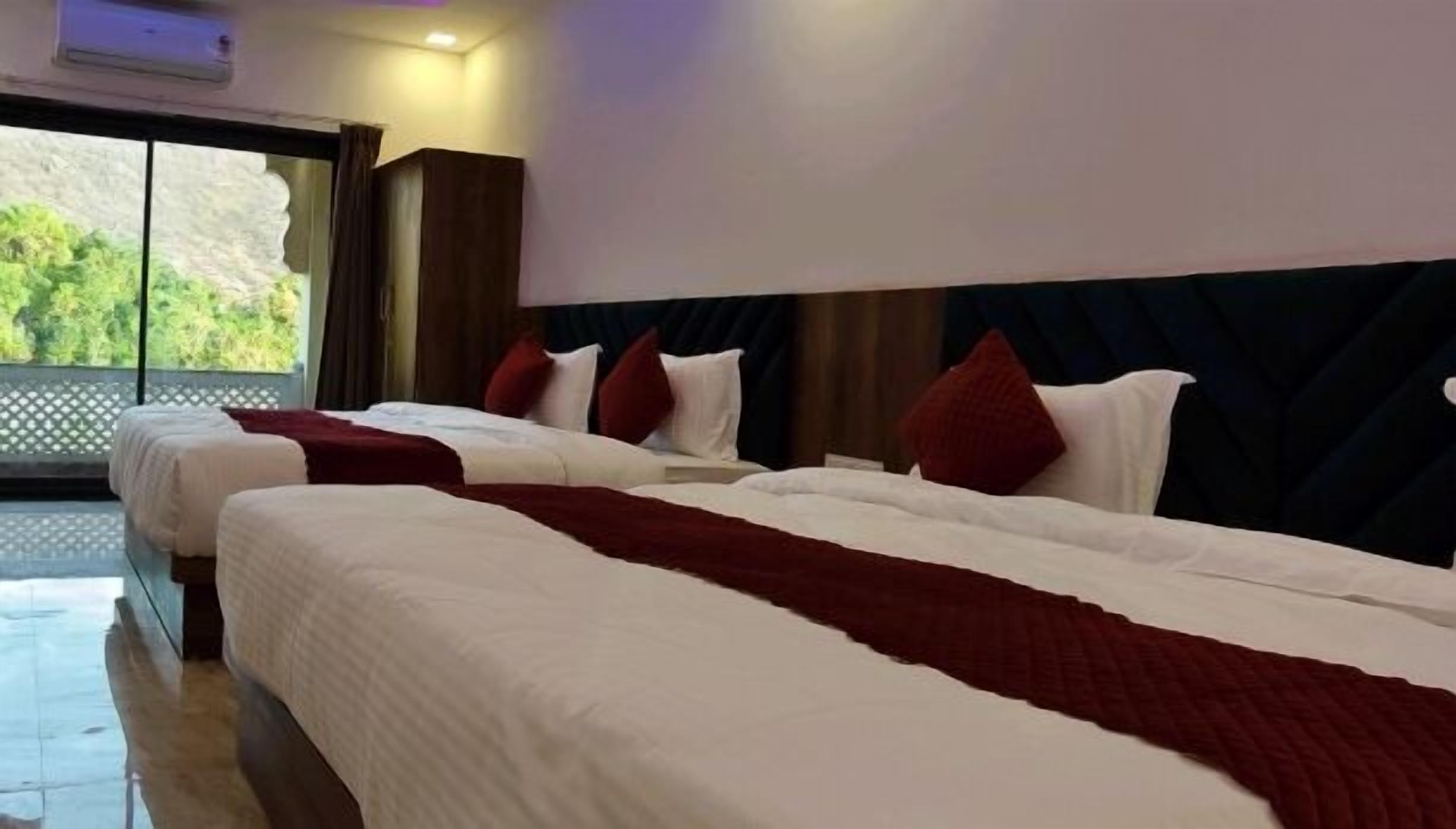 A hotel room with two beds, each with white bedding and red pillows, and a large window offering a view of greenery.