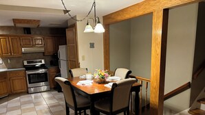 A dining area with a wooden table, four chairs, and a vase of flowers. There is a kitchen in the background with wooden cabinets and appliances.