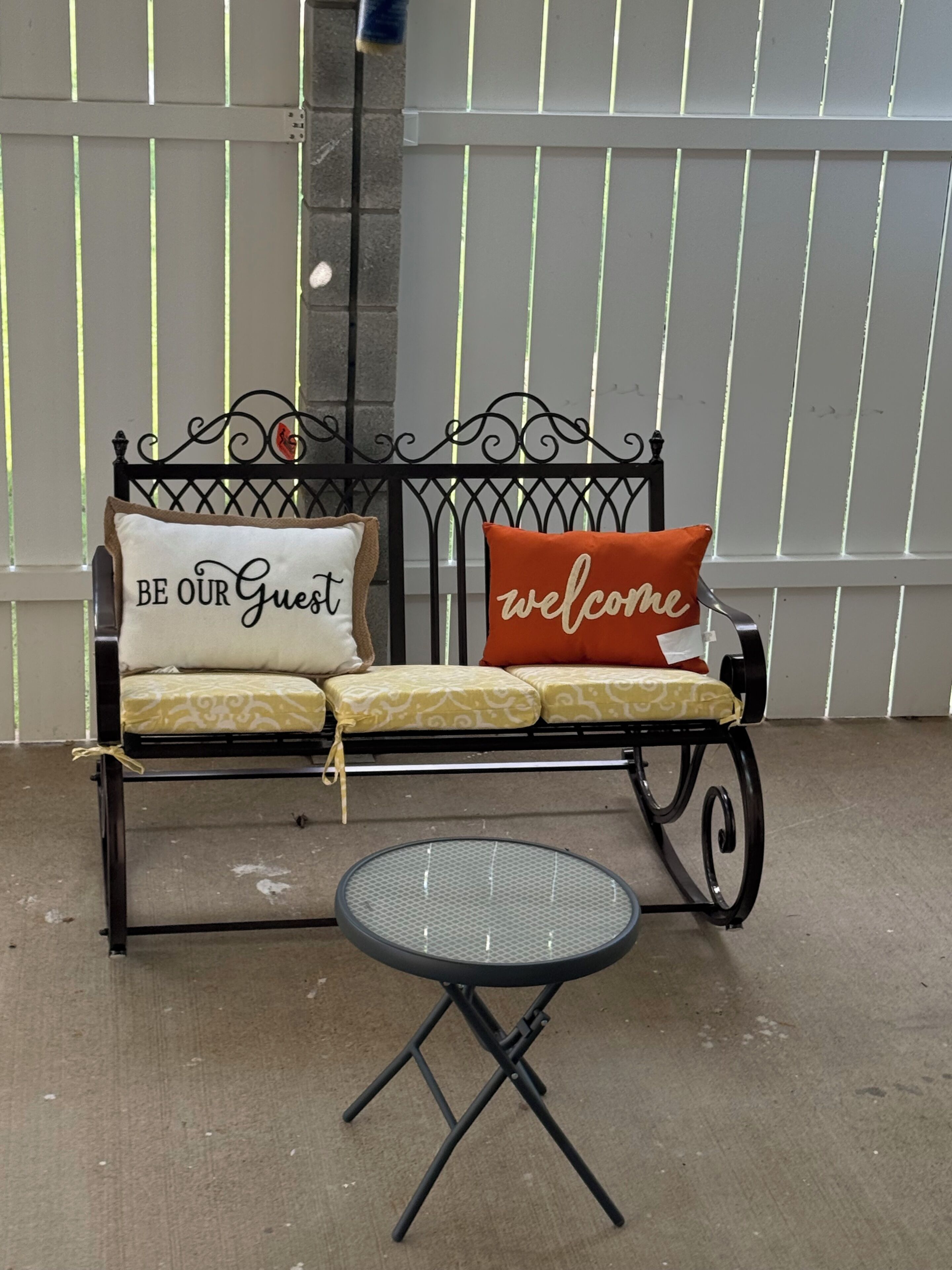 A black metal bench with cushions, one cushion with 'BE OUR Guest' and another with 'welcome'.