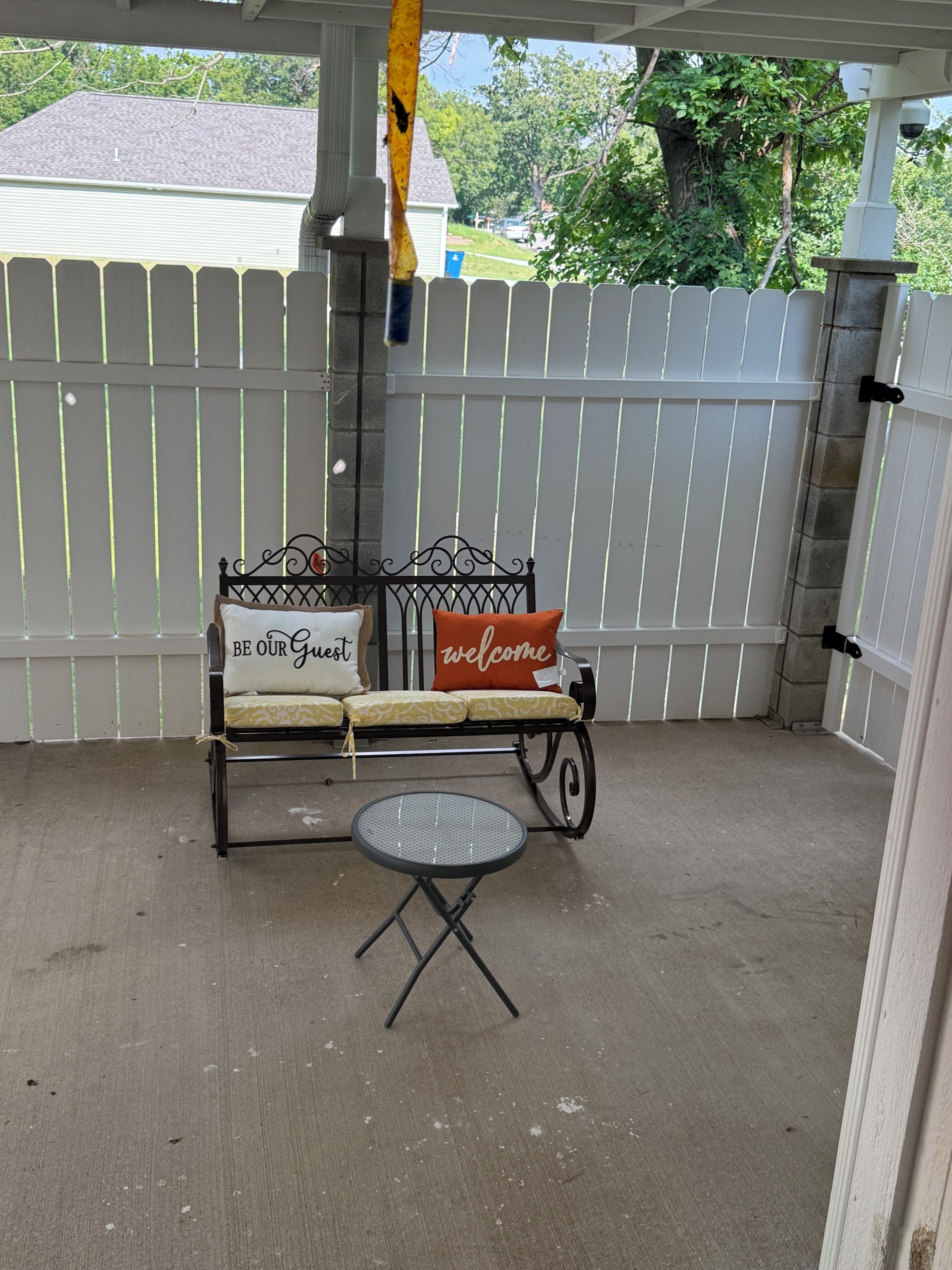 A covered patio area with a metal bench and a small round table, a white fence, and a view of a residential neighborhood.