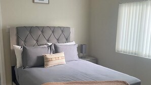 A bedroom with a bed, a headboard, a bedside table, a lamp, a window with blinds, and a picture on the wall.