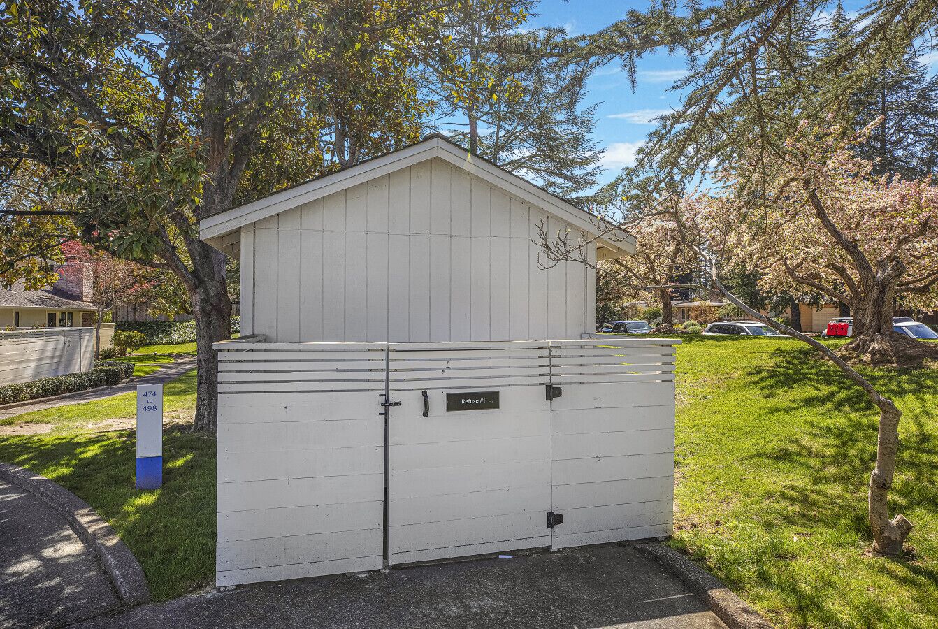 A white shed with a sloped roof and two doors, labeled "Refuse #1" and "Refuse #2," set in a grassy area with trees and a clear sky.