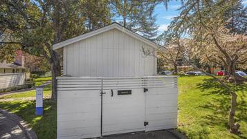 A white shed with a sloped roof and two doors, labeled "Refuse #1" and "Refuse #2," set in a grassy area with trees and a clear sky.