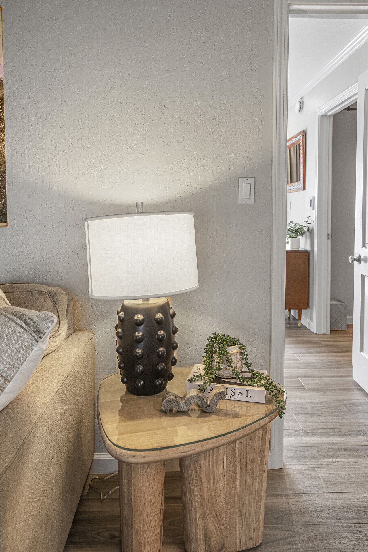 A living room with a beige sofa, a round wooden side table with a lamp, and a small decorative item.