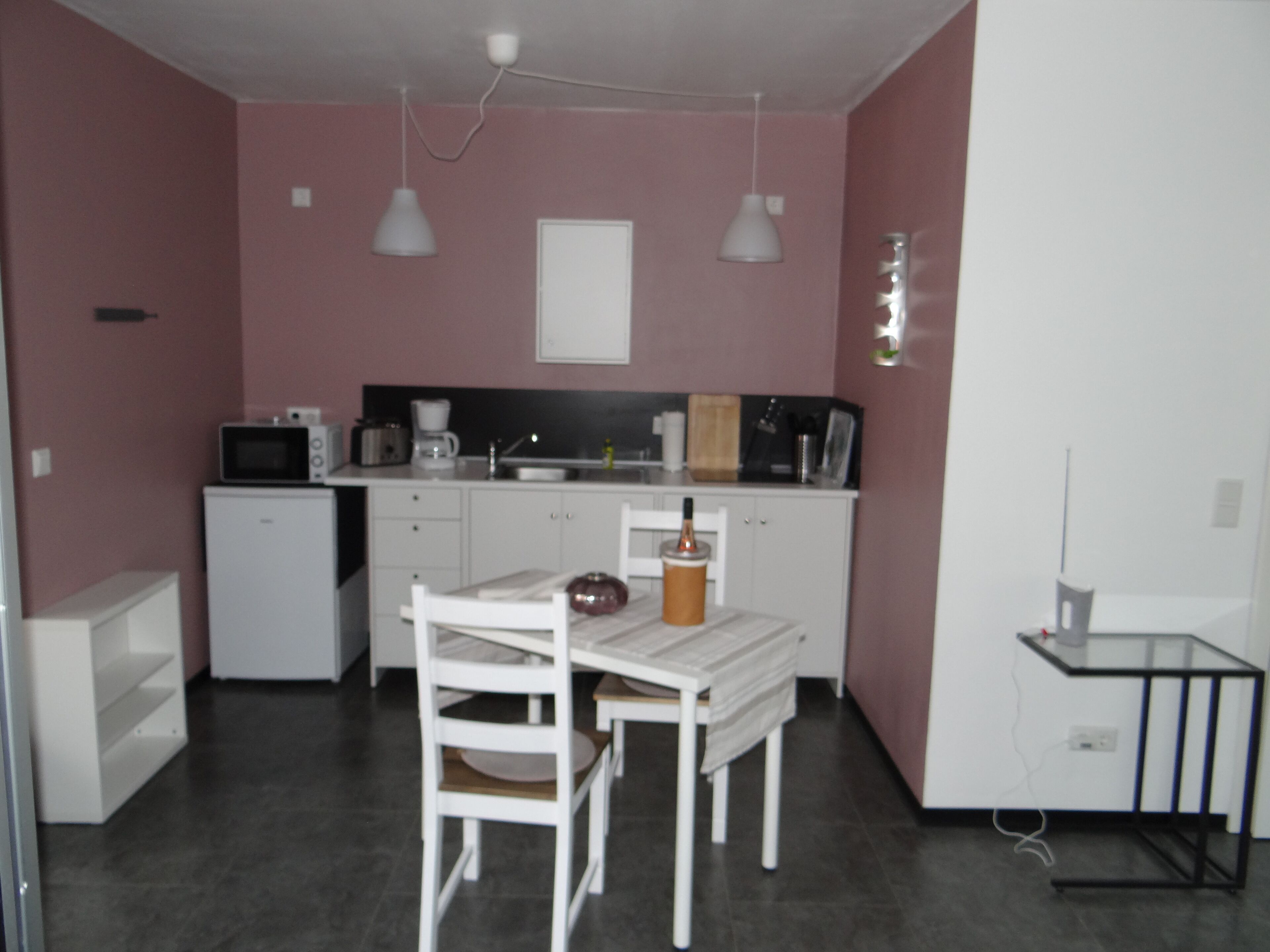 Eine kompakte Küche mit weißen Schränken, einer Mikrowelle, einer Kaffeemaschine und einem kleinen Kühlschrank. Ein Esstisch mit zwei Stühlen und einer einzelnen Kerze steht daneben.