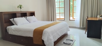 Cama bem arrumada com lençóis brancos e manta marrom, cabeceira de madeira e um pequeno vaso com flores na mesa de cabeceira.