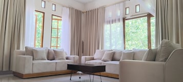 Uma sala de estar moderna com dois sofás, uma mesa de centro e janelas amplas com cortinas translúcidas.